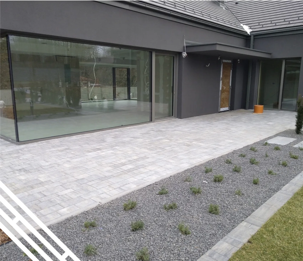 Szara elewacja domu z dużymi przeszkleniami, brukowanym tarasem i pasem ozdobnej roślinności Elewacje domu