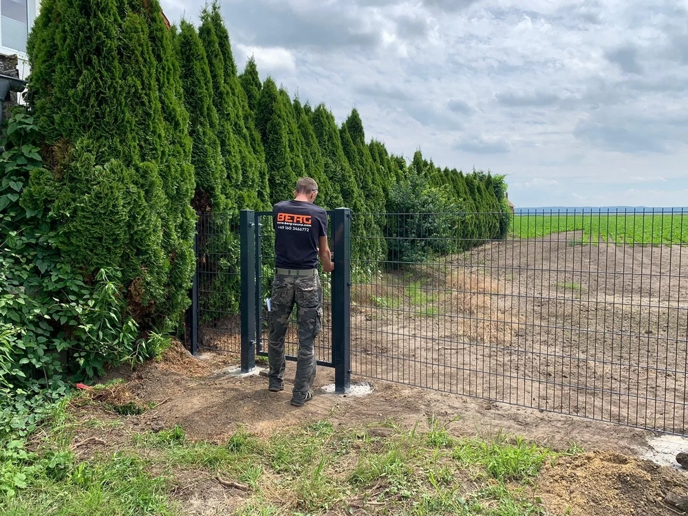 Monteur installiert ein Doppelstabmattenzaun-Tor vor grüner Hecke und Ackerland
