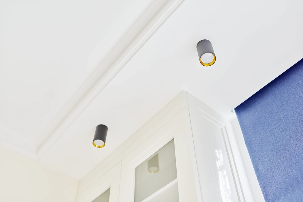 Sufity podwieszane z czarnymi punktowymi oprawami oświetleniowymi, biała zabudowa szafkowa i granatowa zasłona