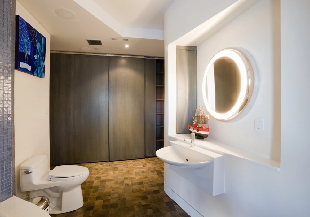 Remont łazienki z nowoczesną umywalką, podświetlanym okrągłym lustrem, toaletą i drewnopodobną posadzką