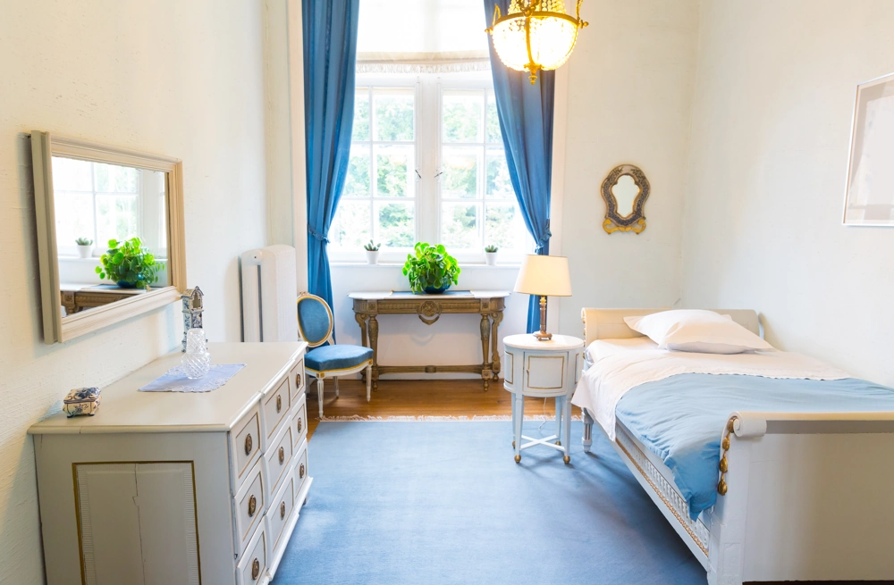 Jasna sypialnia z łóżkiem, komodą, biurkiem, lampą i niebieskimi zasłonami – aranżacja wnętrz